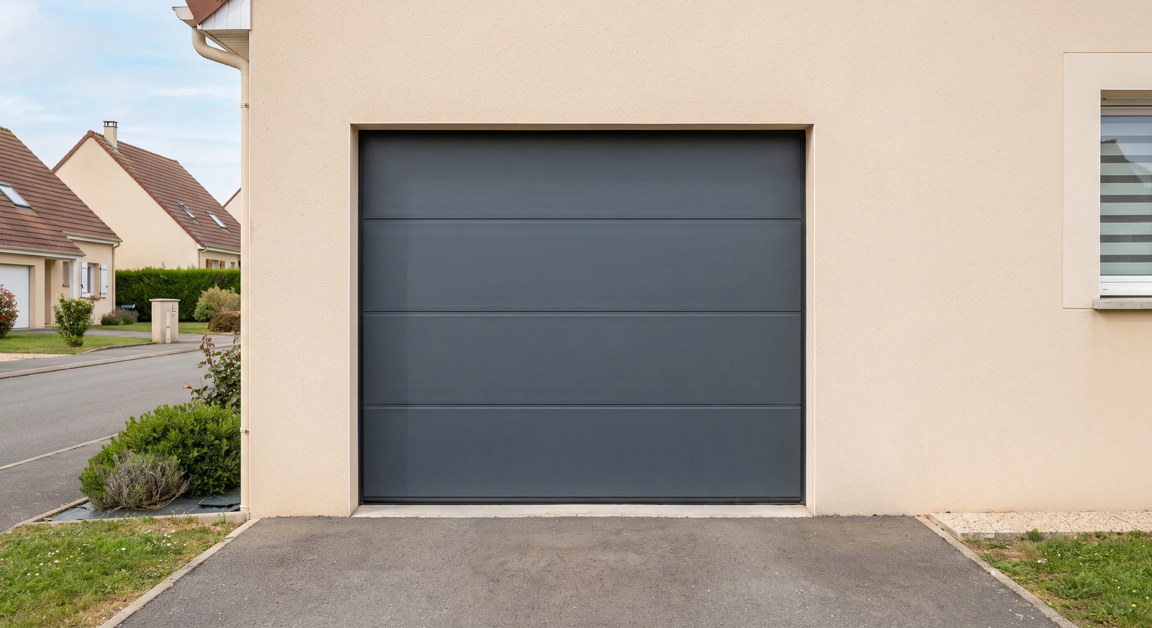 Porte de garage sectionnelle en aluminium gris anthracite avec panneaux horizontaux lisses, vue frontale complète installée sur façade de maison individuelle pavillonnaire, enduit beige, avec allée bitumée en premier plan et environnement résidentiel classique. La porte est fermée, d'aspect professionnel, montrant clairement la menuiserie et ses détails de fixation.