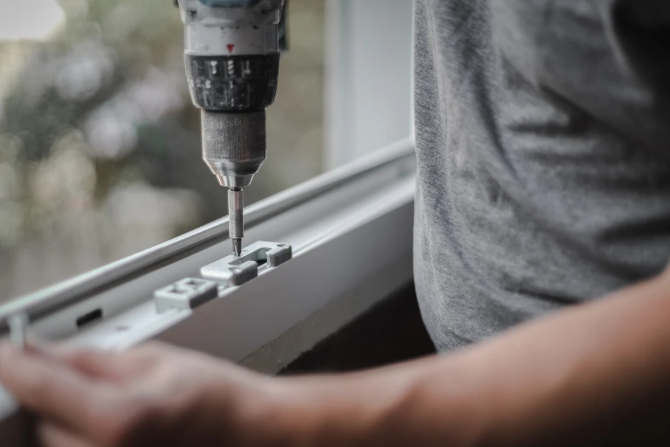 Technicien en tenue de travail professionnelle gris-bleu effectuant un réglage de motorisation sur un portail en aluminium gris anthracite installé en façade extérieure, utilisant un tournevis ou outil de diagnostic, avec en arrière-plan flou une propriété résidentielle. Le technicien porte le gilet ou marquage professionnel de l'entreprise.