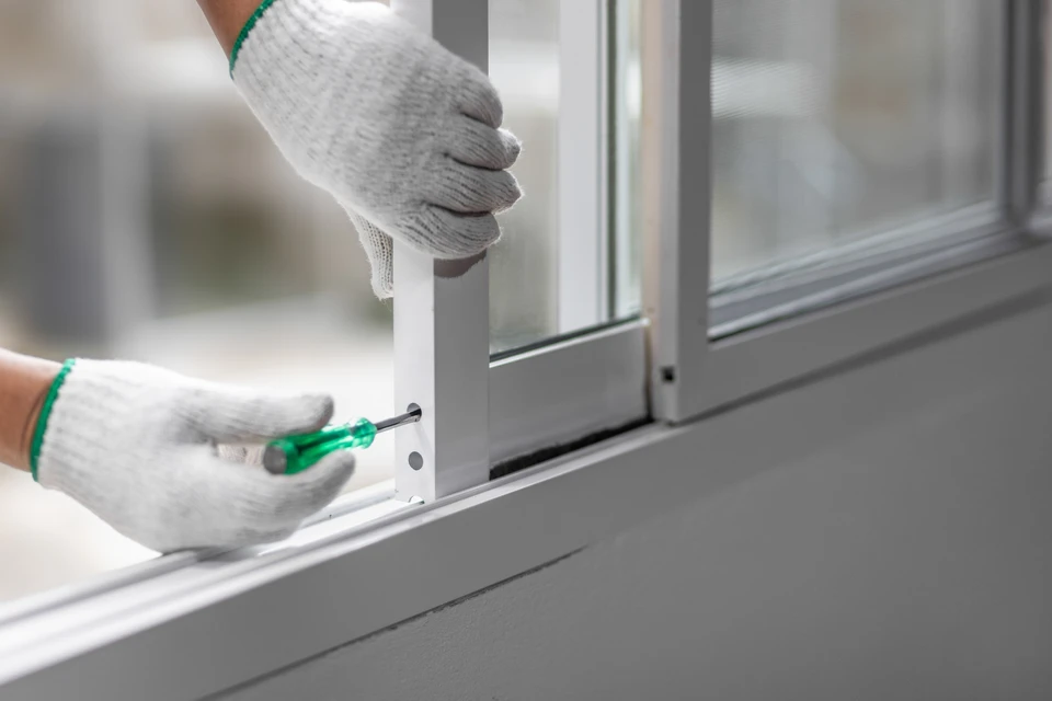 Ouvrier en combinaison bleue et t-shirt vert installe une fenêtre PVC blanc à double vitrage depuis l'intérieur d'un bâtiment
