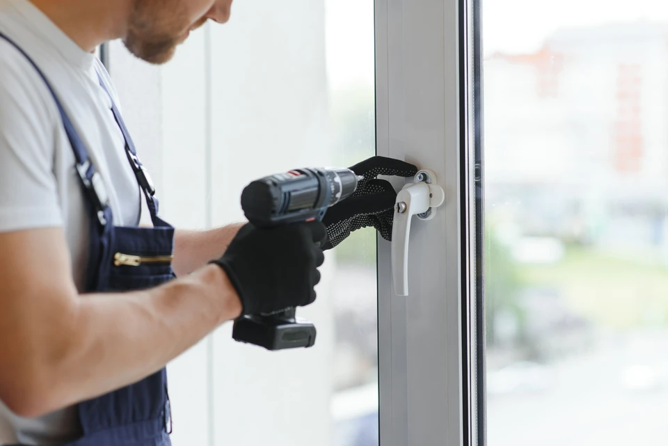 Deux artisans en tenue de travail professionnelle, collaborant ensemble sur l'installation d'un volet roulant en aluminium gris anthracite en façade extérieure. L'un guide l'installation du mécanisme en haut de la fenêtre, l'autre vérifie l'ajustement du tablier. Environnement résidentiel avec enduit de façade clair, fenêtre partiellement visible. Gestes professionnels de précision, concentration partagée.